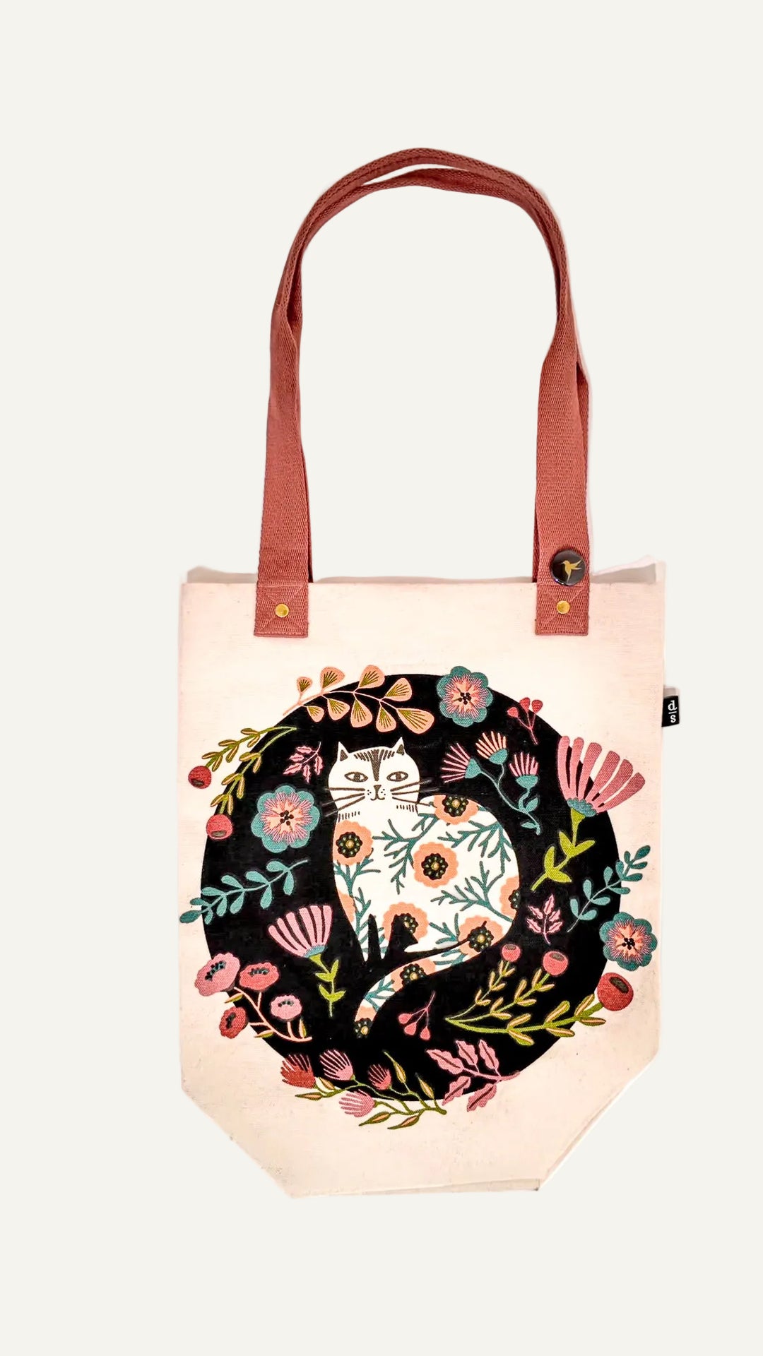 A charming natural off-white canvas tote bag featuring a playful black design of a white cat amidst a vibrant medley of multicolored flowers. Finished with a cheerful pink handle.
